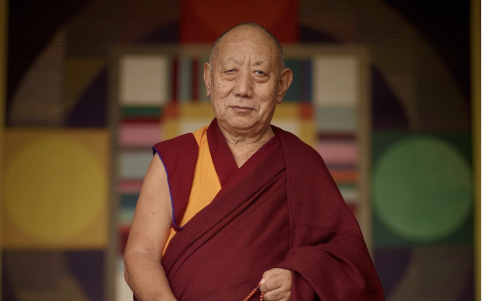 Khensur Rinpoche, a Tibetan monk and master, is standing in front of a mandala image
