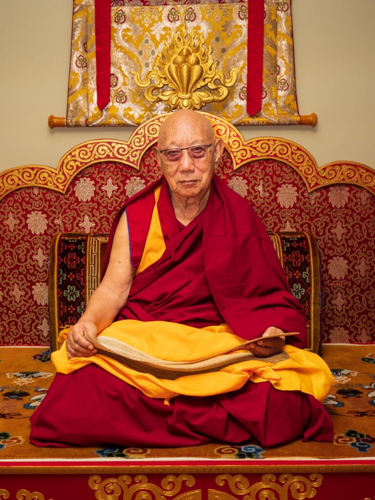 Khensur Rinpoche, a Tibetan monk and master, is sitting on a throne, holding a sacred text.